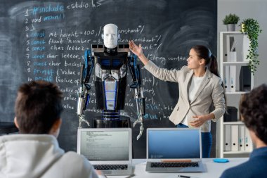 Akıllı kız karatahtanın başında duruyor ve seminerde sınıf arkadaşlarına karakteristik özelliklerini sunarken robotu işaret ediyor.