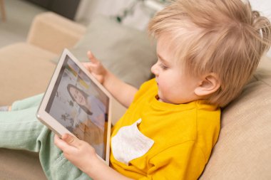 Evdeki karantinada kanepede dinlenirken annesiyle görüntülü konuşma yapan dokunmatik yastıklı mutlu küçük çocuk.