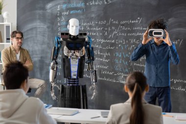 Öğretmen ve genç sınıf arkadaşları vr kulaklıklı bir öğrenciye bakıyor. Derste robotun karatahtaya karşı sunumunu yapıyorlar.