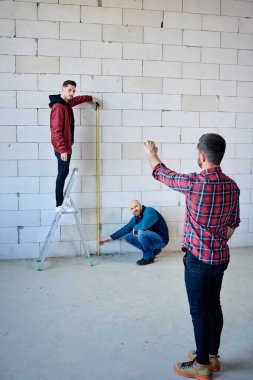 Duvardaki cetveli düzenleyen genç mühendisler meslektaşlarına bakar ve ölçüm sırasında onun talimatlarını dinlerler.