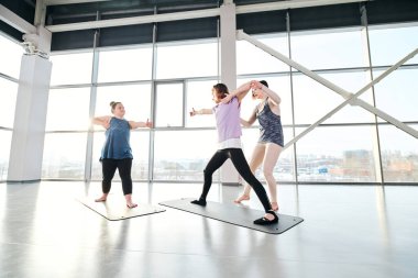 Spor kıyafetli iki genç kadın yoga egzersizlerinden birini yaparken minderlerin üzerinde duruyor ve eğitmenleri antrenman sırasında onlara yardım ediyor.