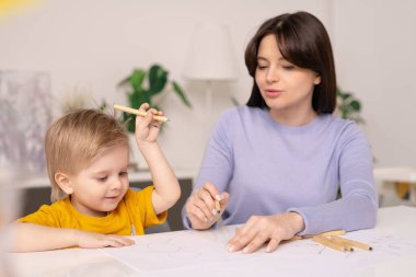 Pastel boyalı kalemle resim çizerken elini masanın üstüne koyan sevimli küçük çocuk ve genç annesi ona yardım ediyor.