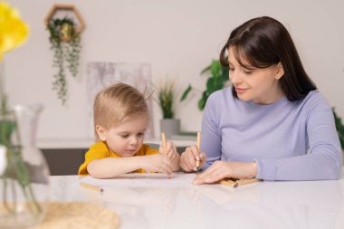 Güzel genç anne ve sevimli küçük oğlu, karantina döneminde evde kalırken renkli boya kalemleriyle resimler çiziyorlar.