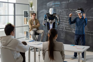 VR kulaklıklı ergen öğrenci sınıf arkadaşlarının önünde tahtada duruyor ve robotun yeteneklerini gösteriyor.