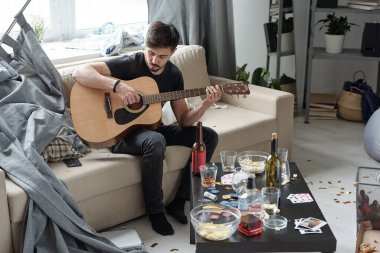 Genç adam perdesiyle kanepede oturuyor ve partiden sonra dağınık bir dairede gitar çalıyor. Alkollü bir masa ve parçalarla oynuyor.