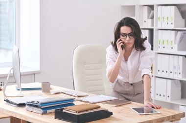 Genç bir sekreter cep telefonuyla bir müşteriyle konuşuyor. Belgeler, tablet ve bilgisayarla masanın üzerine eğiliyor.