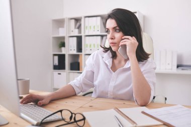 Ciddi genç sekreter ya da ofis yöneticisi bilgisayar ekranının önünde oturuyor, telefonla konuşuyor ve verilerle çalışıyor.