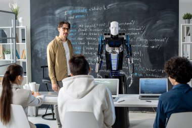 Genç öğretmen izleyicilerin önünde tahtada duruyor ve ders sırasında bir grup öğrenciye robot sunumu yapıyor.