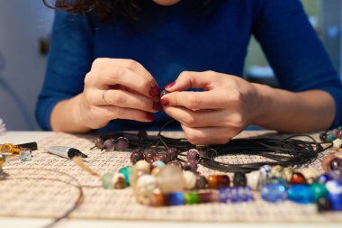 İş yerinde oturan ve atölyede kolye yaparken plastik ipliğe cam boncuklar takan genç yaratıcı kadının elleri.