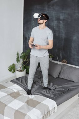 Yatağında sakallı yakışıklı genç adam sanal gerçeklik gözlükleriyle oyun oynarken joystick kullanıyor.