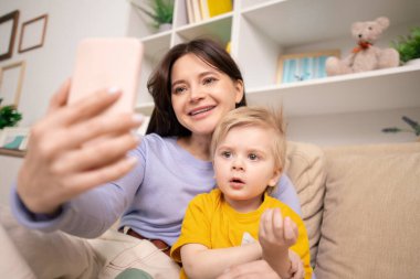 Genç, gülümseyen bir kadın ve sevimli oğlu kanepede oturup akıllı telefondaki kameraya bakarak ev ortamında selfie çekiyorlar.