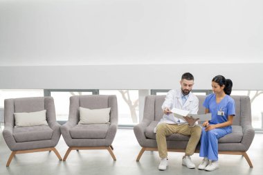 Sakallı genç klinisyen, modern kliniklerin bekleme salonunda asistanıyla konuşurken hastalarından birinin reçetesini ya da kartını gösteriyor.