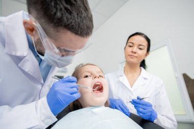 Ağzı açık, dişçi muayenesi sırasında diş kliniğinde koltukta oturan dişçisinin aynası ile konuşan sevimli küçük bir kız.