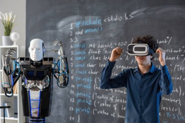 VR kulaklıklı çağdaş öğrenci karatahta ile el kaldırırken derste robot yeteneklerini sunuyor.