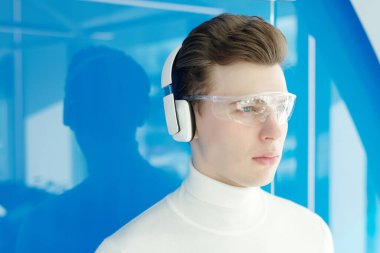 Kulaklıklar ve akıllı gözlüklerle gelecekteki teknolojilerin genç ve kendinden emin geliştiricisi modern çağda mavi duvarın yanında duruyor.