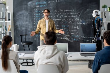Modern teknoloji kursunun kendine güvenen öğretmeni seyircilerin önünde tahtaya karşı duruyor ve ders puanlarını anlatıyor.