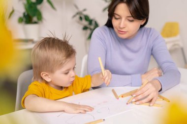 Memnun genç anne masada oturuyor ve oğlunun renkli kalemlerle resim çizmesine yardım ediyor.