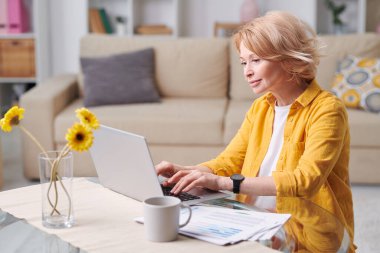 Normal giyinen mutlu sarışın kadın dizüstü bilgisayarın önünde oturuyor ve karantina sırasında ev ortamında uzaktan çalışıyor.