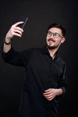 Gözlüklü yakışıklı genç adam siyah tişörtlü selfie çekiyor. Akıllı telefonuyla izole edilmiş karanlık arka plana karşı.