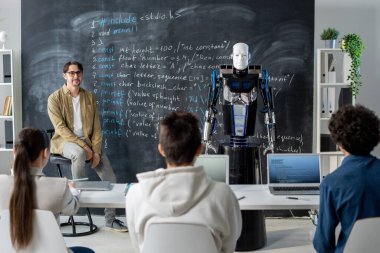 Bir grup genç öğrenci tahtada duran robota bakıyor ve teknik özelliklerini seminerde öğretmene anlatıyor.