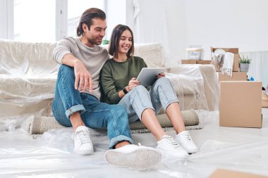 Mutlu, genç, sevgi dolu bir çift yeni evlerinin ya da evlerinin oturma odasında oturuyorlar ve online mobilyaların arasında kayıyorlar.