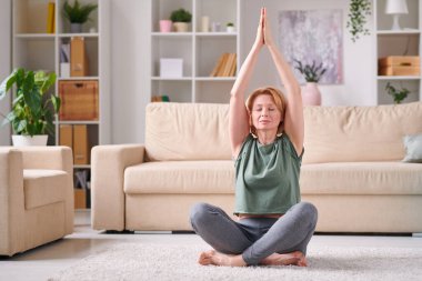 Aktif kıyafetli sağlıklı olgun bir kadın karantinada evde otururken ellerini birleştirip nilüfer çiçeği pozisyonunda oturuyor.