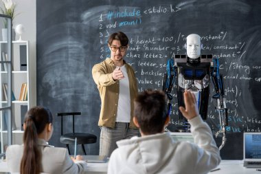 Çağdaş öğretmen, tahtada duran robotun özellikleri hakkında soru sormak isteyen öğrencilerden birini işaret ediyor.