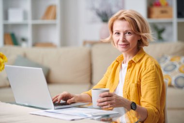 Successful mature businesswoman with mug of tea looking at you while sitting by desk in front of laptop at home during quarantine clipart