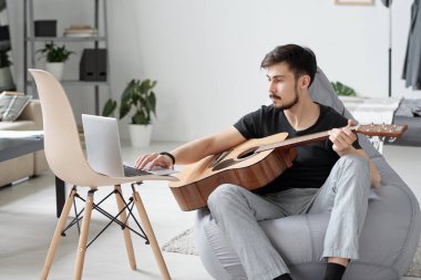 Ciddi bir genç adam karantina sırasında evde gitar çalmayı öğrenirken bilgisayarını kullanıyor.