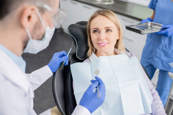 Молодая улыбающаяся блондинка, пациентка стоматологических клиник, сидящая в кресле и смотрящая на своего стоматолога с крюком и зеркалом перед осмотром
