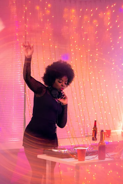 在迪斯科舞会上 年轻的非洲裔女主持人一边站在桌子旁一边 一边用耳机听音乐 — 图库照片