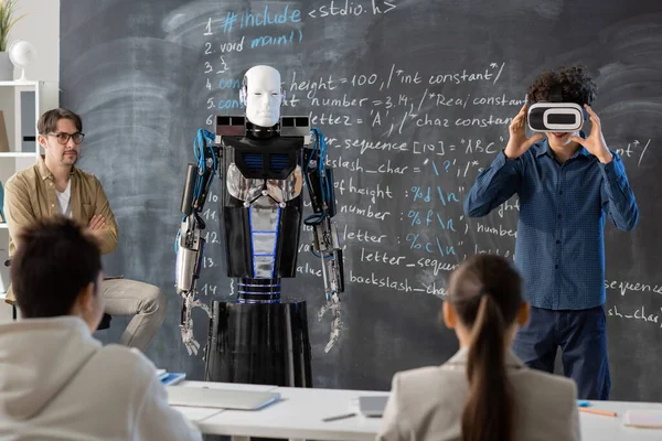 Öğretmen ve genç sınıf arkadaşları vr kulaklıklı bir öğrenciye bakıyor. Derste robotun karatahtaya karşı sunumunu yapıyorlar.