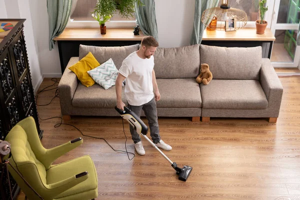 Young man in casualwear doing domestic work while cleaning floor in living-room with vacuum cleaner between armchair and couch