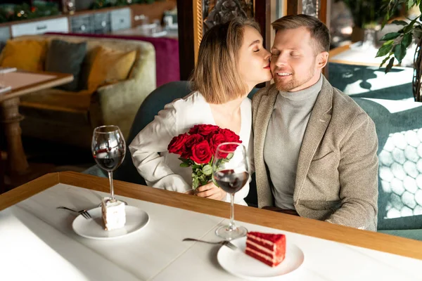 年轻而快乐的女人 戴着华丽的红玫瑰花束 双双坐在餐桌前亲吻男朋友的脸颊 — 图库照片
