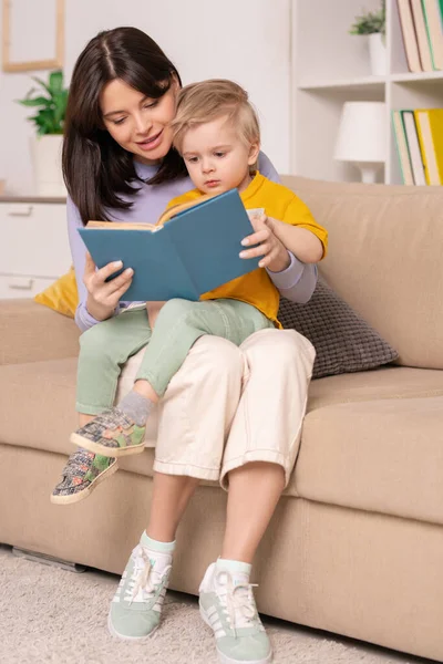 快乐的年轻母亲坐在沙发上 抱着小儿子 阅读着带着照片的儿童读物 — 图库照片