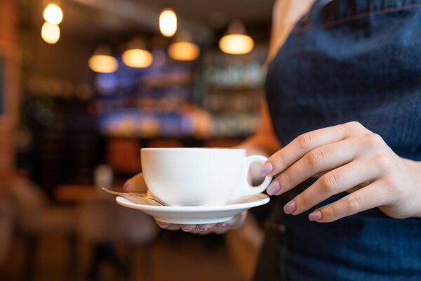 Hand of young waitress with cup of tea or coffee carrying it to one of clients while working in contemporary restaurant or cafe