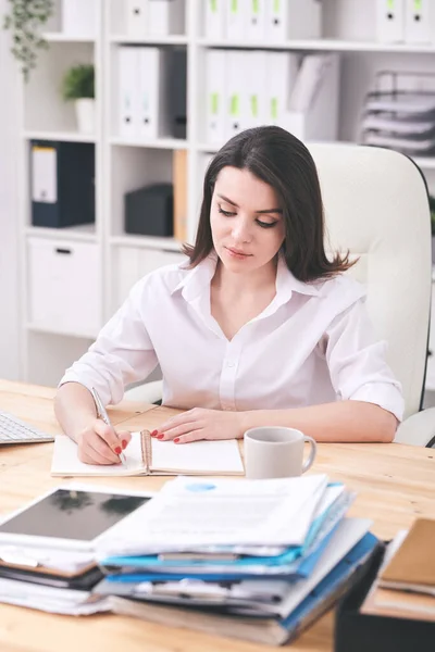 若い真剣な女性の白いシャツに座っているアームチェアで机と棚の背景にノートブックで作業計画を立てる — ストック写真