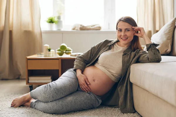 Retrato Uma Jovem Grávida Sorridente Sentada Chão Apoiada Sofá Enquanto — Fotografia de Stock