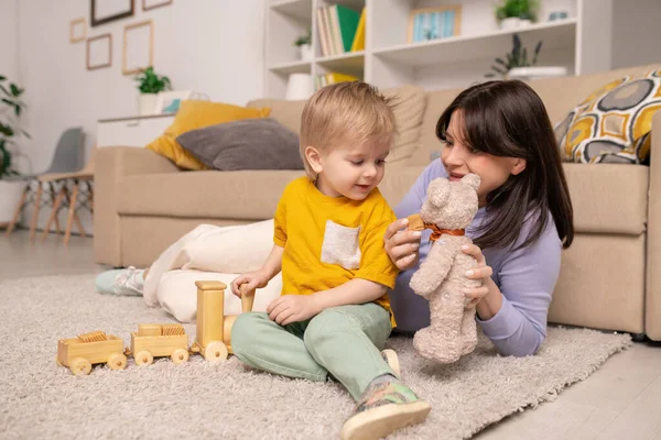 快乐的年轻妈妈躺在地毯上 把玩具熊展示给儿子看 而他们则在家里一起玩玩具 — 图库照片