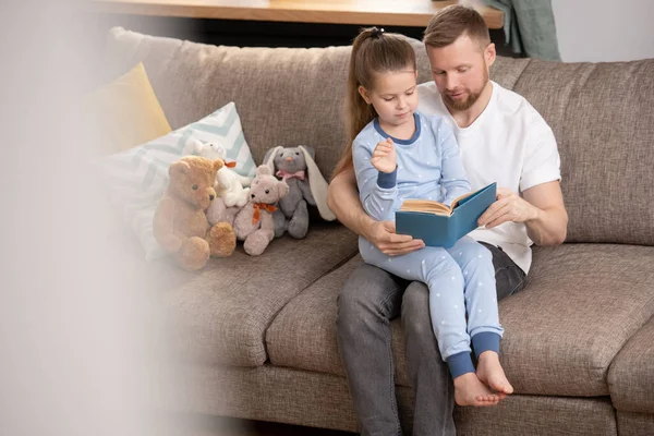 穿着睡衣的漂亮女孩坐在父亲的膝上 闲暇时或周末一起在沙发上看童话 — 图库照片