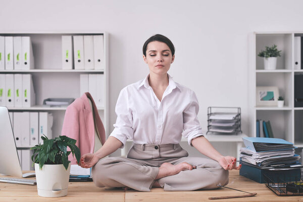 Pretty young barefoot businesswoman in formalwear sitting in pose of lotus on desk in office while trying to concentrate or relax