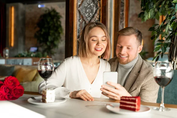 年轻的一对笑容可亲的情侣坐在高档餐厅的餐桌旁 一边喝酒一边吃蛋糕 一边用智能手机看什么 — 图库照片
