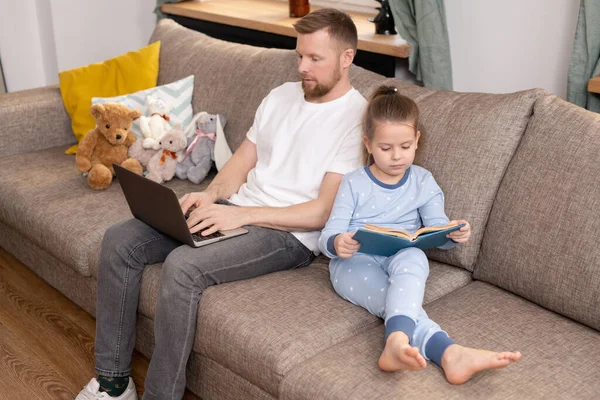 在检疫期间 穿着蓝色睡衣的小女孩坐在沙发上 阅读童话 而她的父亲则拿着笔记本电脑远程工作 — 图库照片