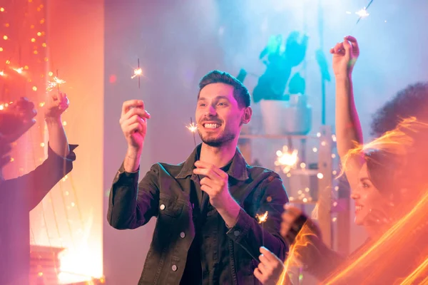 新年のパーティーで友人と楽しい時間を過ごしながら 彼の手の中に輝くベンガルライトを見て歯の笑顔で幸せな若い男 — ストック写真