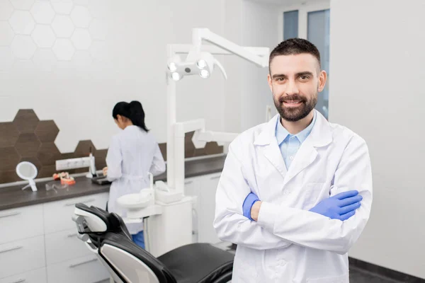 年轻快乐的 留着胡子的专业牙医 戴着手套 站在摄像机前 身后站着他的助手 — 图库照片