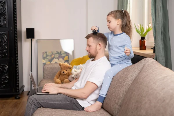 在隔离期间 漂亮的孩子们一边在客厅里坐在沙发上 一边在笔记本电脑前梳理着她父亲的头发 — 图库照片