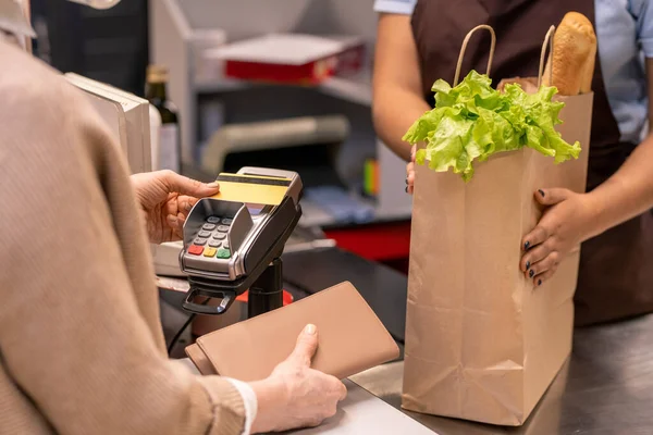 クレジットカードで商品を支払う成熟した女性のバイヤーの前で新鮮な食料品と紙袋に若いレジ係の手 — ストック写真