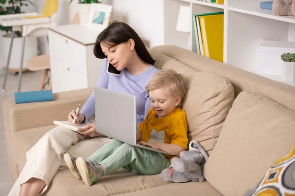 快乐可爱的小男孩坐在沙发上 坐在年轻妈妈旁边 一边用智能手机聊天 一边在笔记本上记笔记 一边玩笔记本电脑 — 图库照片