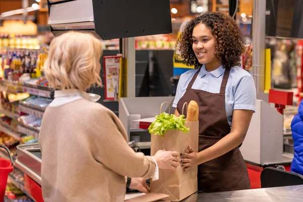 スーパーのレジに立っている間 パンや新鮮な食料品と成熟した女性の紙袋を与えるかなり若いレジ係 — ストック写真