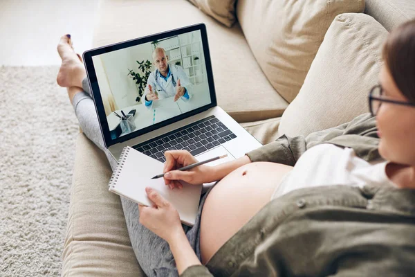 怀孕妇女躺在沙发上 在网上与医生讨论怀孕问题时做笔记 — 图库照片